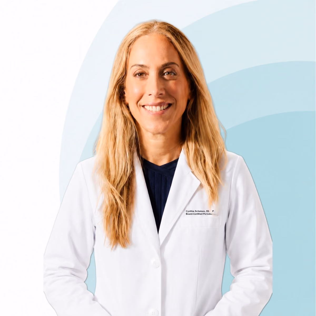 Smiling woman with long blonde hair wearing a white lab coat over a dark top, standing against a light blue and white background.