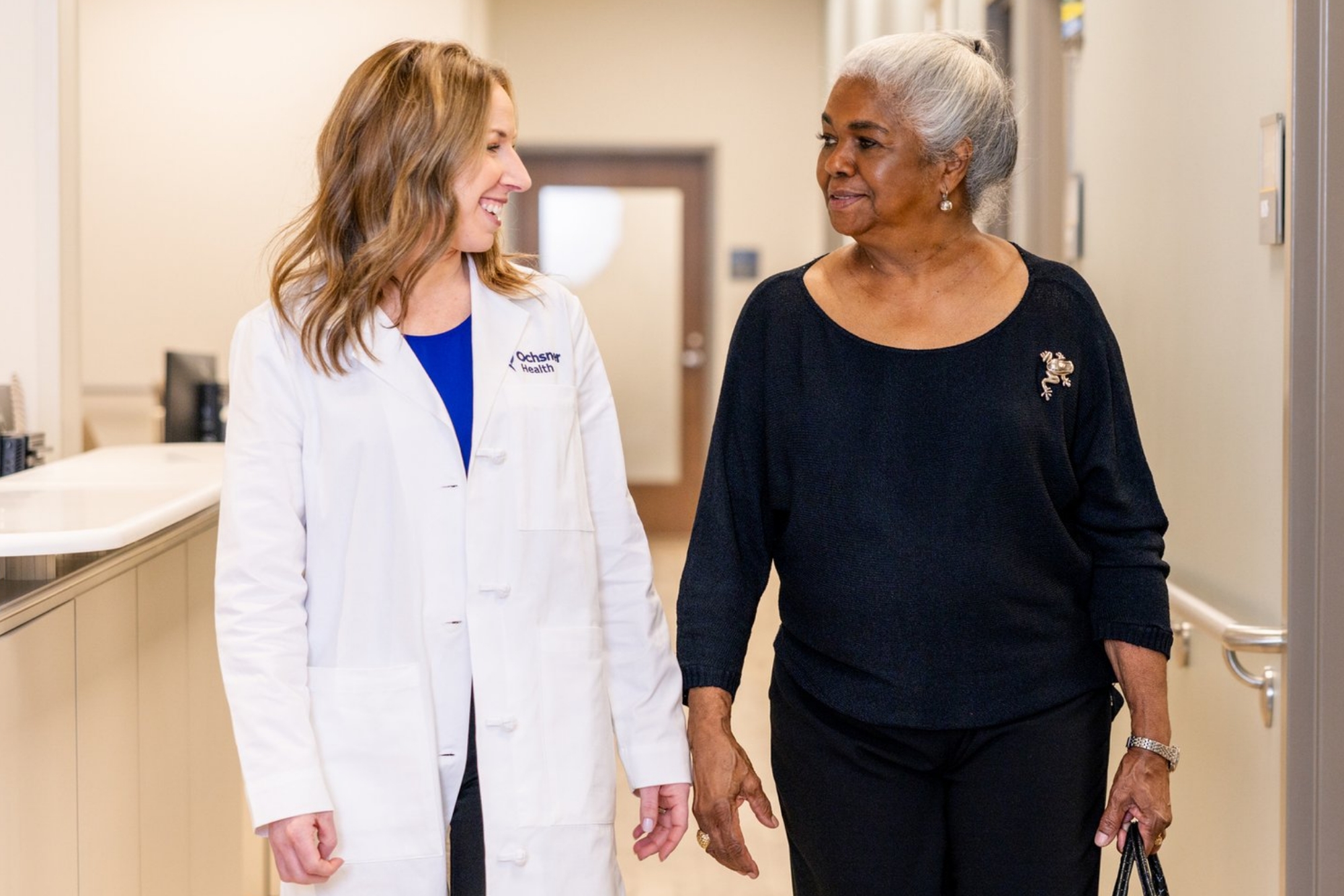 Doctor in a white coat walking down a clinic hallway and smiling while talking with an older woman holding a purse, suggesting a friendly patient-provider interaction.