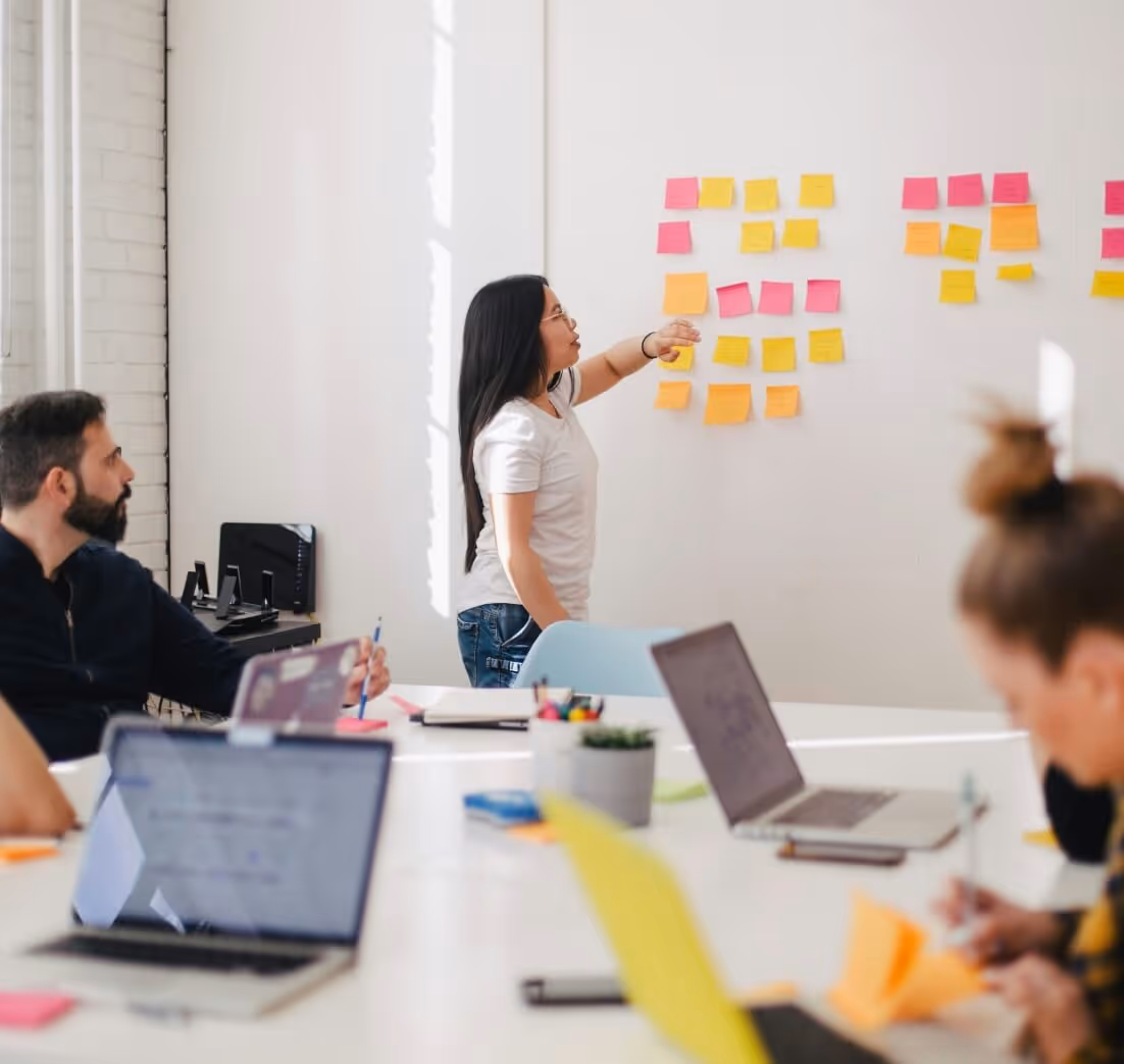 Image of a female pointing towards a group of sticky notes on a whiteboard with a body of team members in the foreground