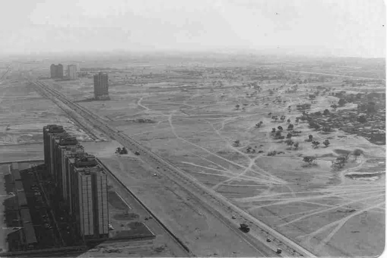 Alt text: A historical black and white photograph showcasing the early stages of urban development in a desert landscape. Sparse high-rise buildings stand along a main road that dissects the barren terrain dotted with small shrubs. 
