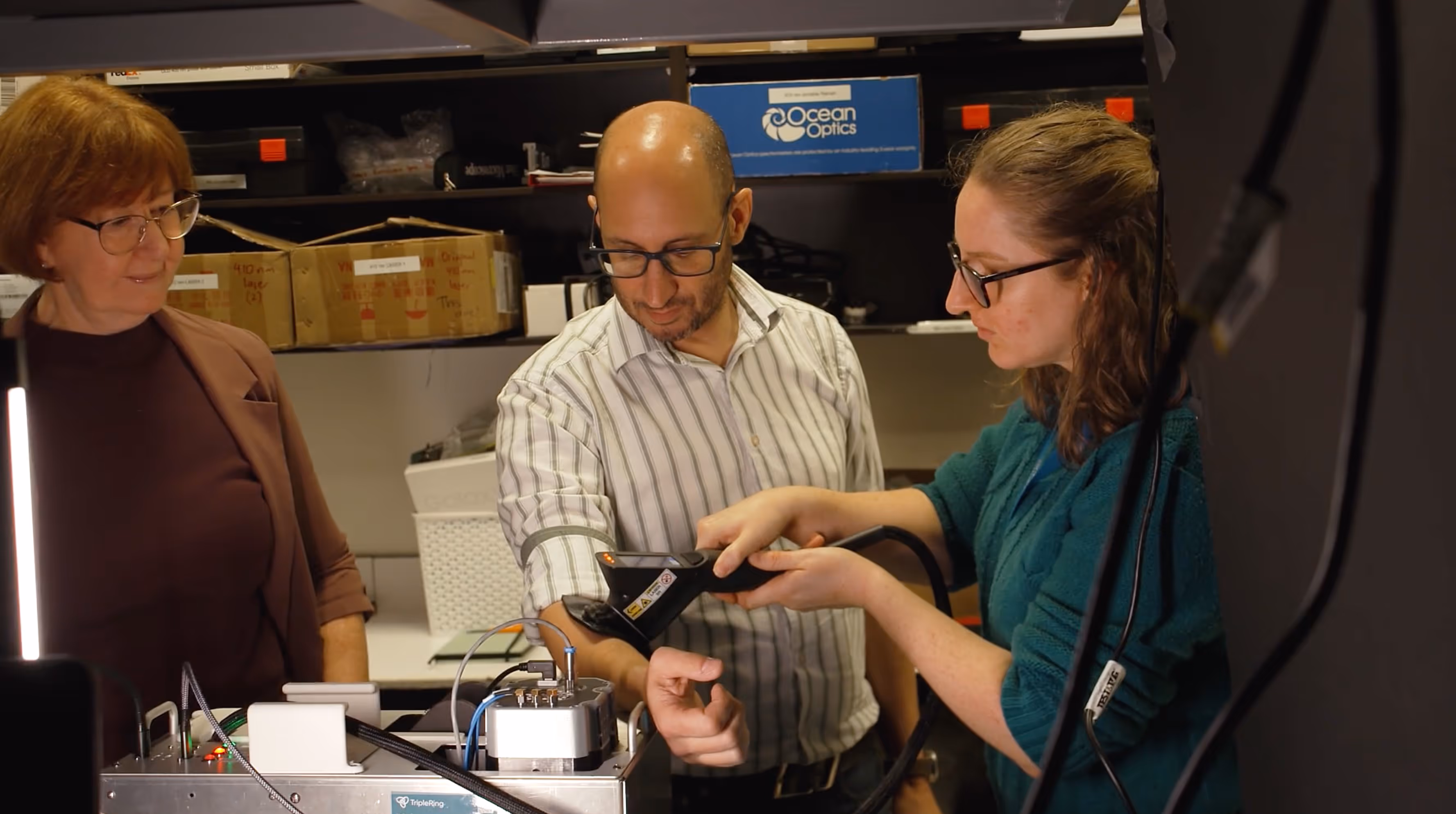 A woman uses a handheld Luminoma device to scan a man's forearm while another woman watches in a laboratory setting.