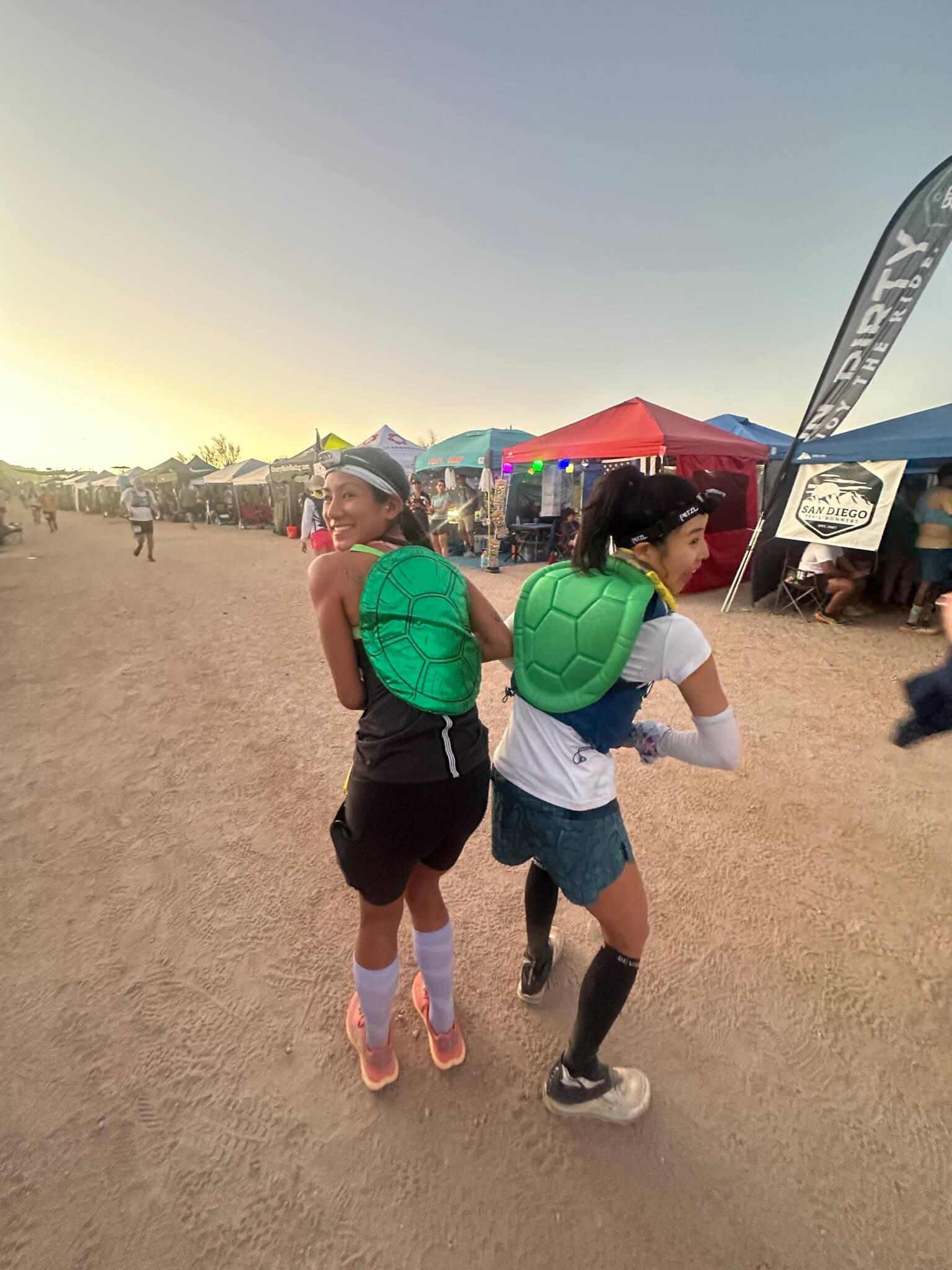 Two women wearing green turtle shells are walking on a beach.