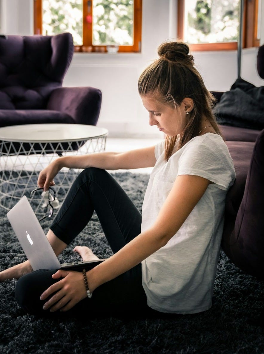 woman in white t-shirt and black leggings sitting on area rug while facing silver MacBook