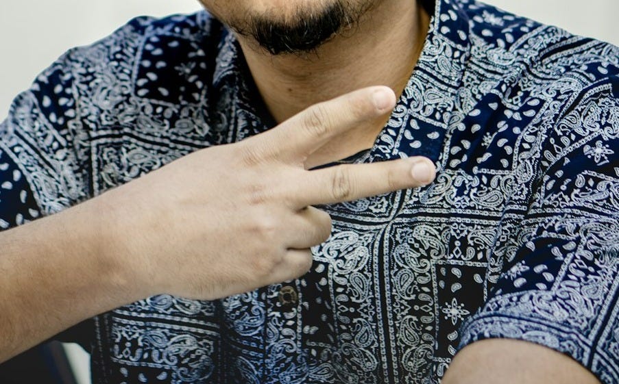 A man with a mustache making a peace sign in a patterned shirt