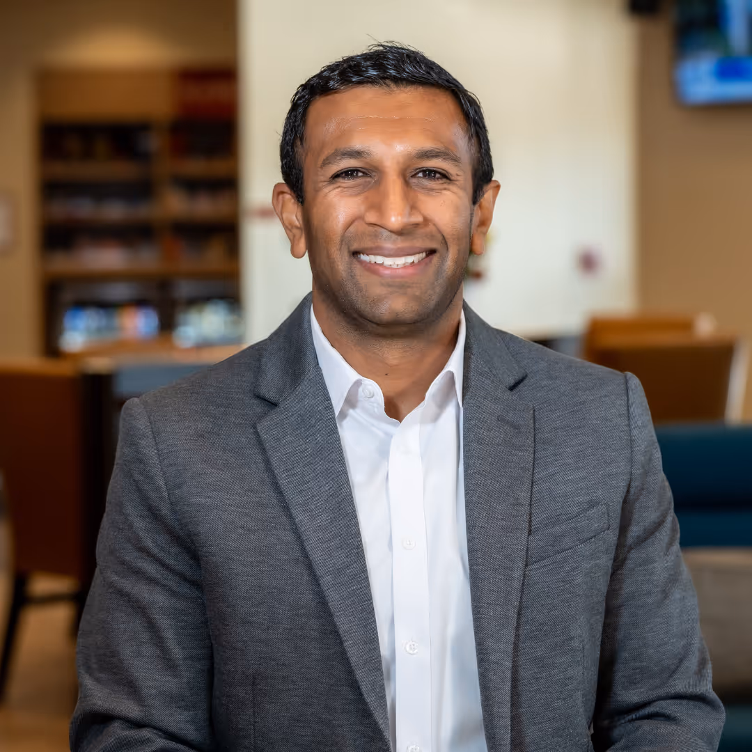 Bhavesh Lad, smiling in a gray blazer and white dress shirt, seated in a warmly lit, blurred background setting.