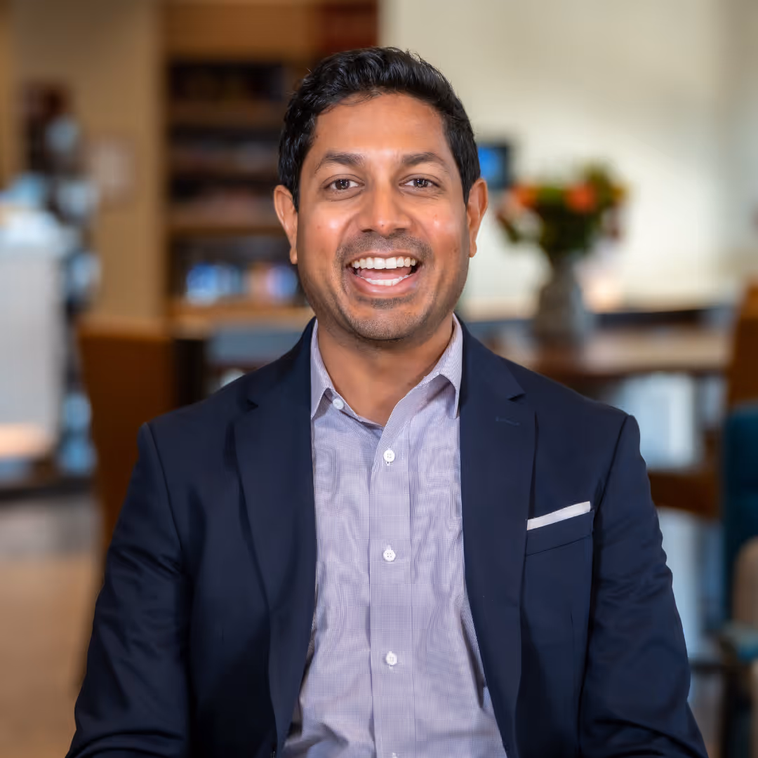 Amit Patel, smiling in a dark blazer and light dress shirt, seated in a warmly lit, blurred background setting.