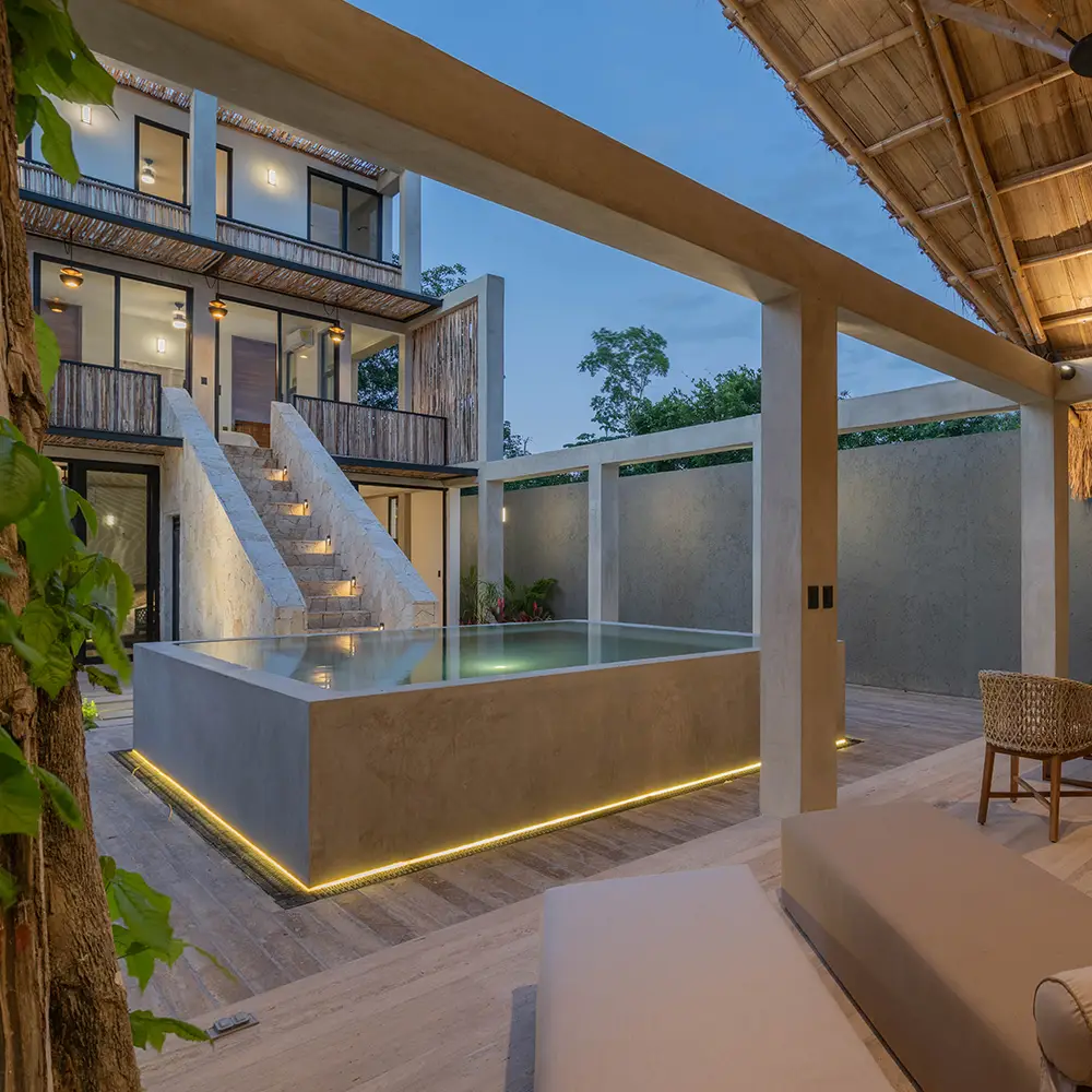 Piscina elevada con iluminación LED en una villa moderna con escaleras de piedra y mobiliario exterior bajo techo de bambú.