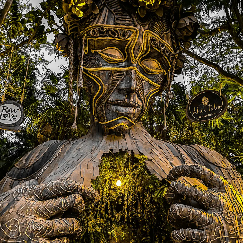 Wooden sculpture of a giant human figure with carved details and yellow lights on the chest, surrounded by vegetation.