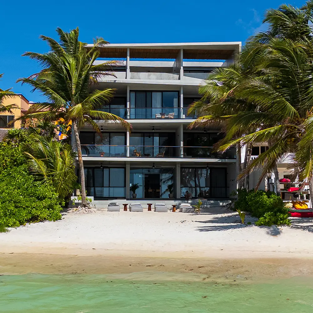 Edificio moderno de tres pisos frente a una playa con palmeras y sillas en la arena bajo un cielo azul despejado.
