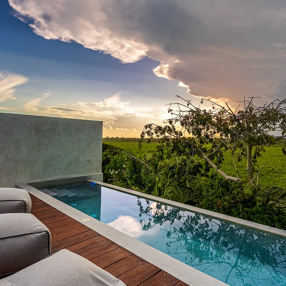 Piscina al aire libre con agua reflejando el cielo nublado al atardecer, junto a sillas acolchonadas y una vista de árboles y campo verde.