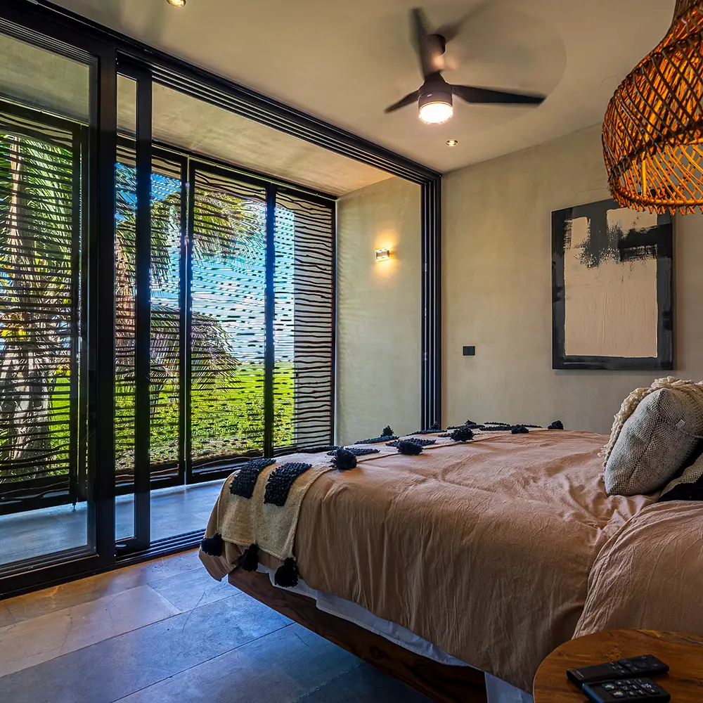 Modern bedroom with queen-size bed, ceiling fan on and sliding window with a view of palm trees and nature.