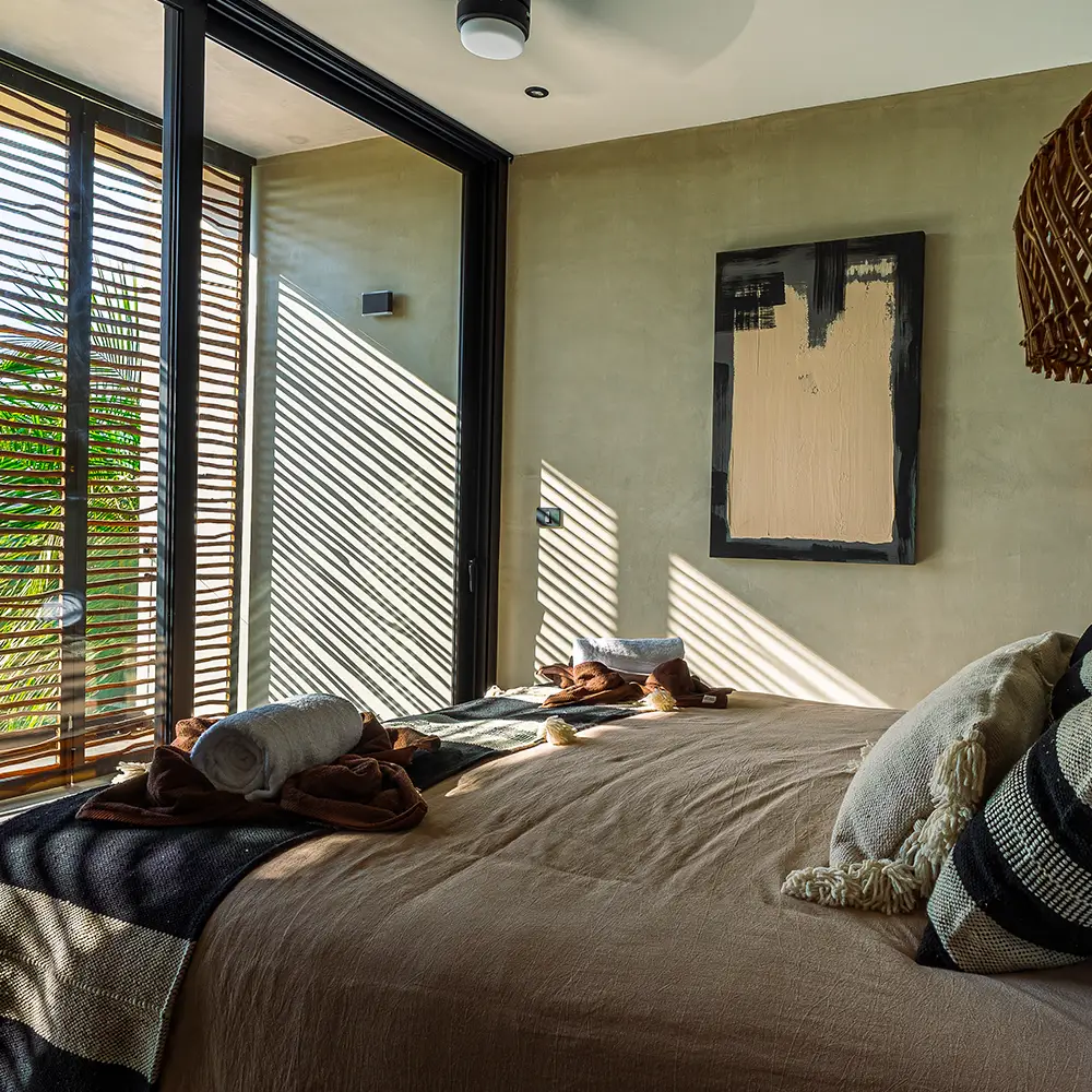 Dormitorio moderno con cama grande, ventilador de techo encendido y ventana corrediza con vista a palmeras y naturaleza.