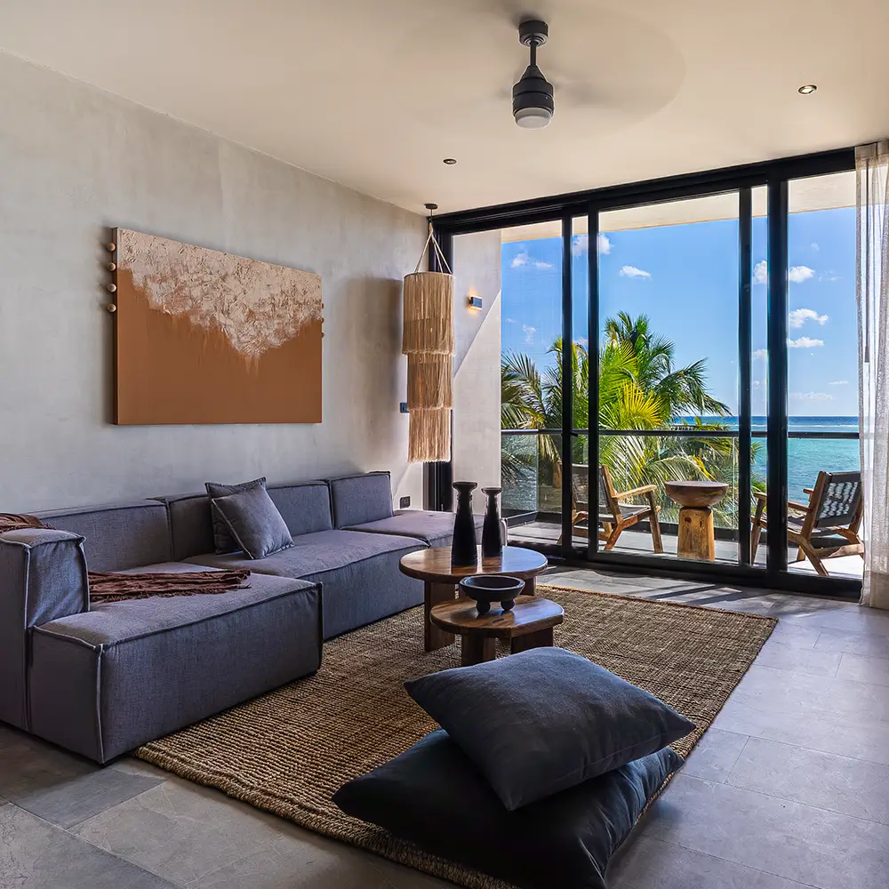 Sofás beige con cojines al lado de una piscina frente a una playa con mar turquesa y palmeras bajo un cielo azul con nubes.