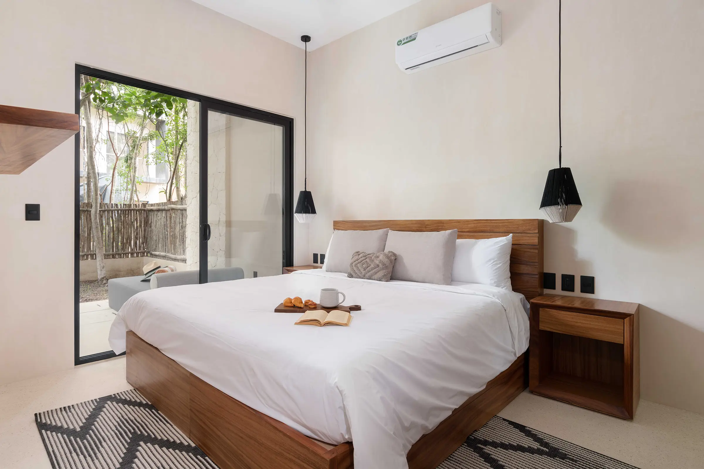 Dormitorio moderno con cama de madera, ropa blanca, lámparas colgantes negras y puerta corrediza a patio.