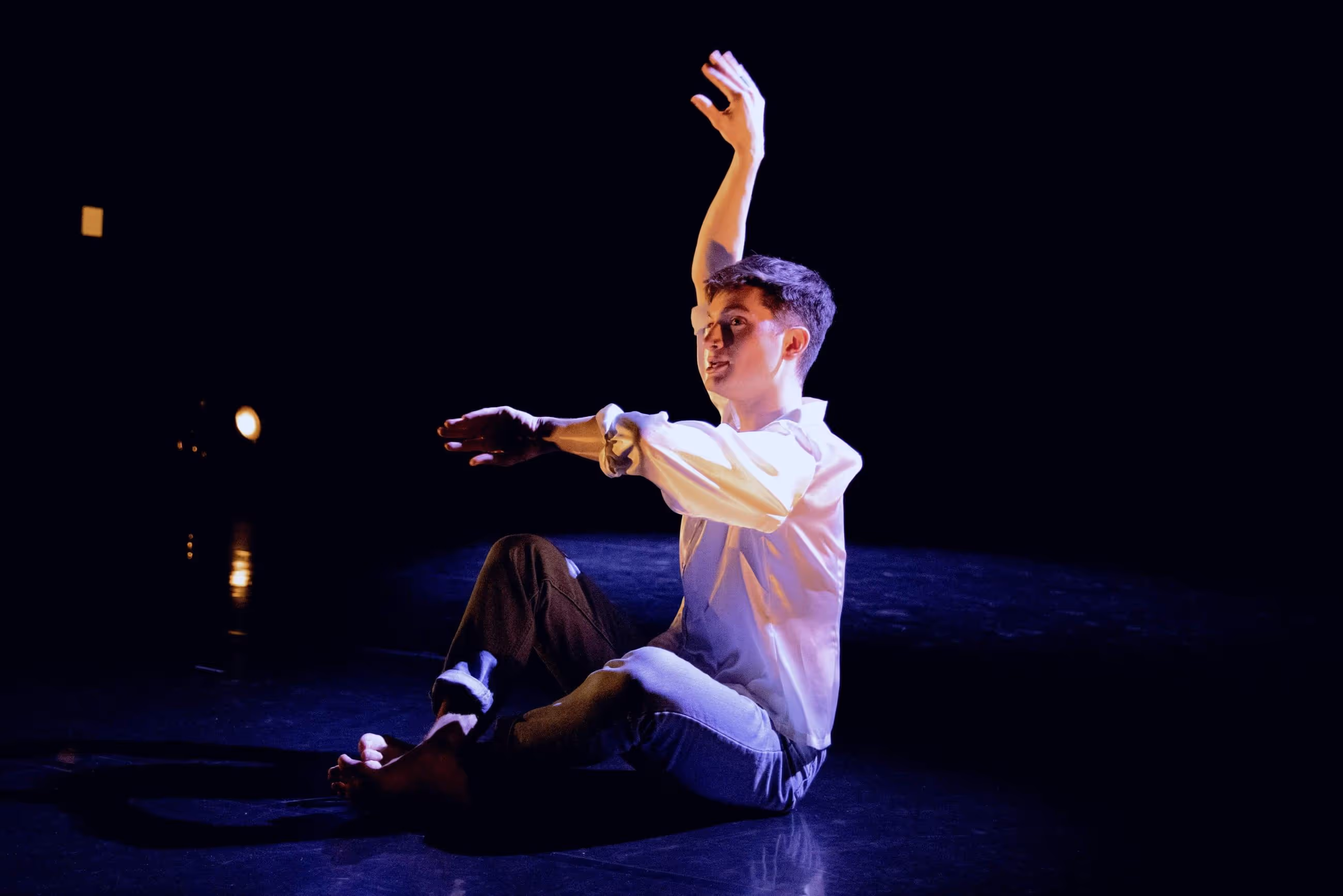 A young person is sitting on a stage, their arms half raised above their body.