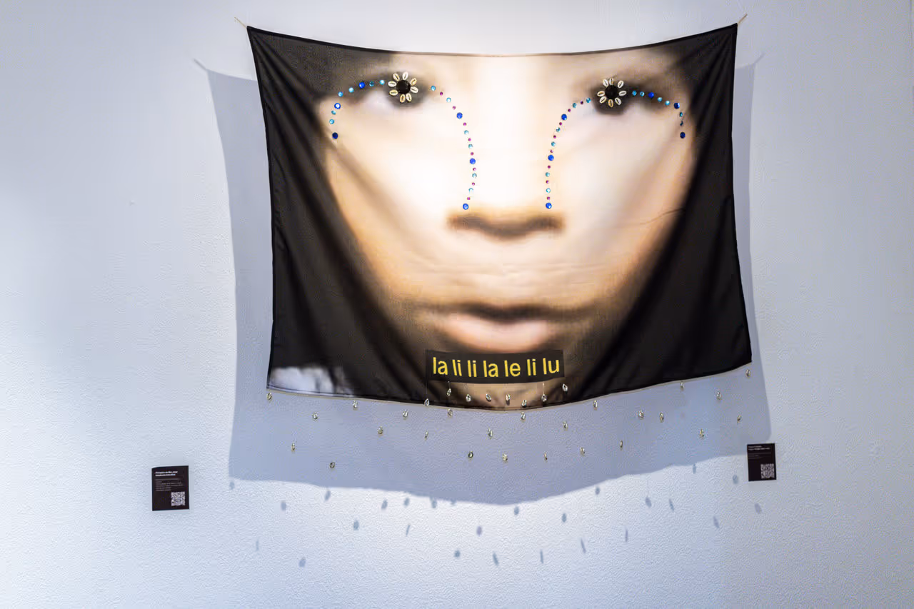 A photograph of an artwork on a white wall. It is a printed fabric with an obscure face and featured beads.