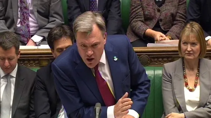 A photograph of Ed Balls speaking in the UK House of Commons. He is a middle-aged man with light skin and greying hair. He is wearing a navy suit with a red tie. He is in the middle of speaking and is leaning against a podium. In the background are his political colleagues sitting on green-upholstered benches.