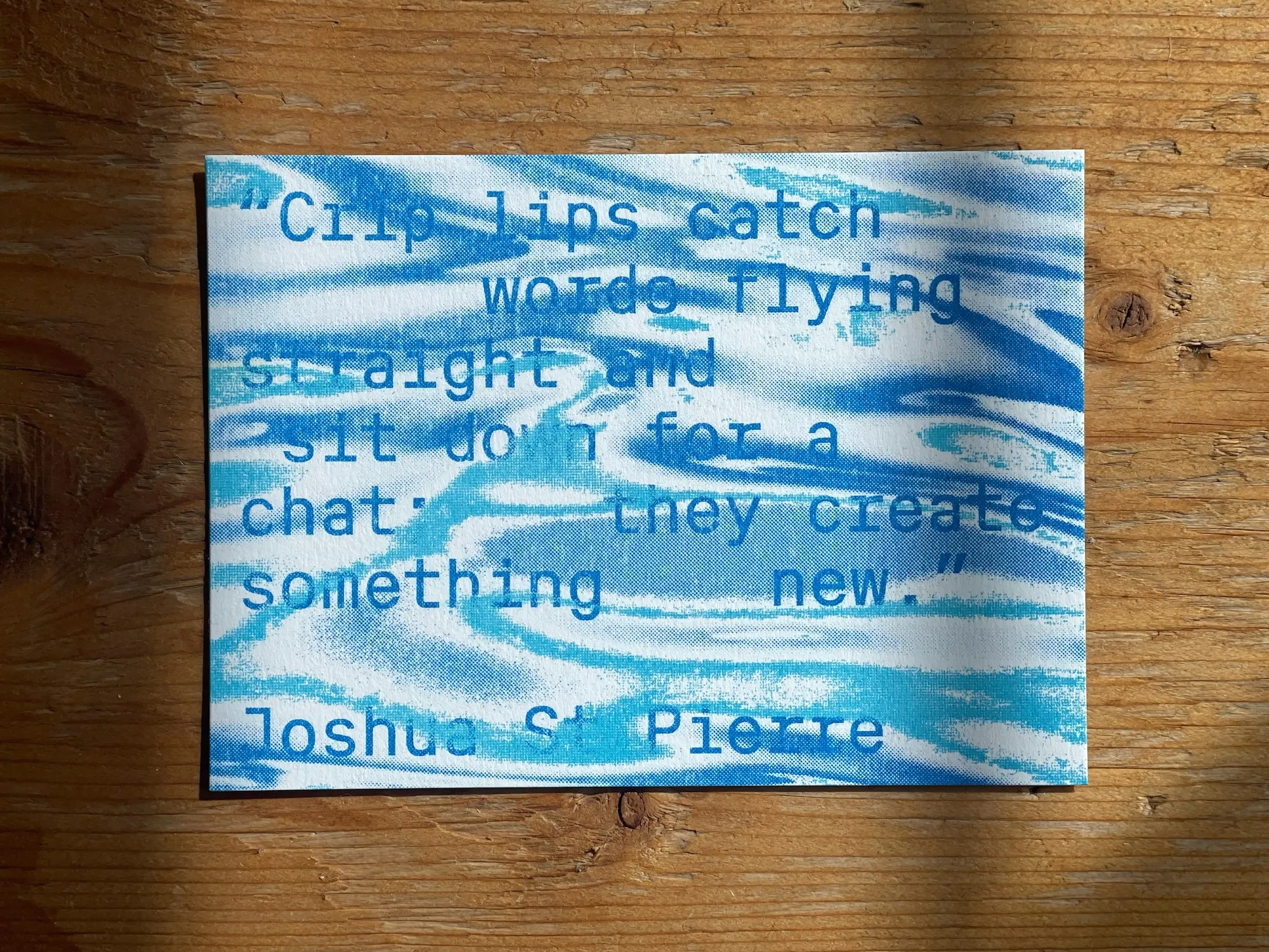 A photo of a postcard on a wooden table. It reads: "Crip lips catch words flying straight and sit down for a chat; they create something new." The quote comes from Joshua St Pierre, whose name is featured on the bottom of the postcard. The font repeats and stretches some letters to reflect a sense of stammering. The text is blue, with blue and aqua waves of colouring the background.