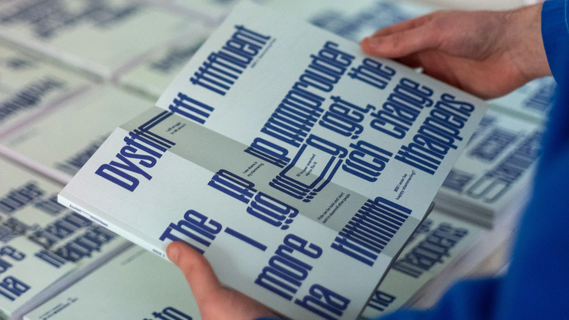 A photo of a person holding Dysfluent magazine issue 2. The front cover is folded outward, revealing a flurry of dark blue, repeated typography that visually represents stammering. The background of the front cover is a light teal. Stacks of the magazine are visible in the background of the phootgraph.