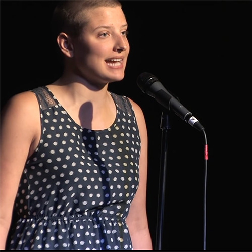 A photograph of Erin Schick speaking in front of a microphone. They have light skin, short hair, and they are wearing a navy and white polka dot garment. The background is totally black.