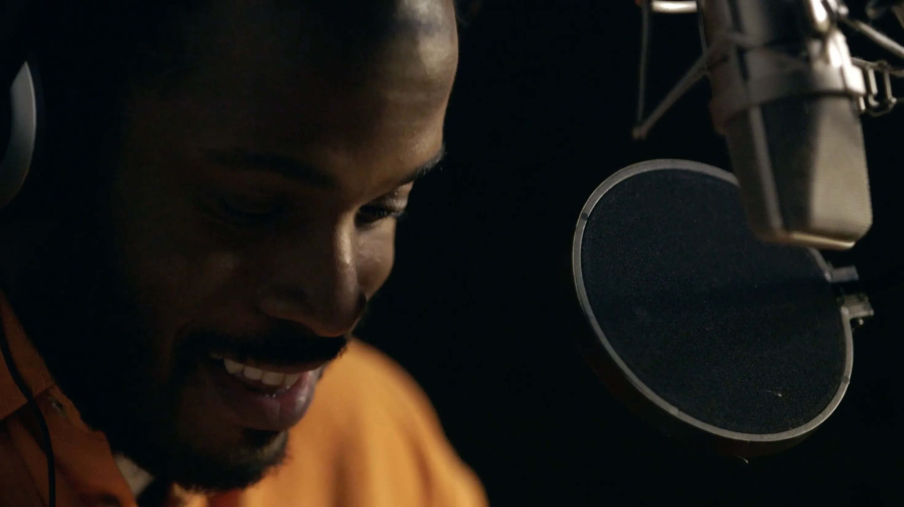 A close-up photograph of a person speaking into a boom microphone. They have dark skin and are smiling, their face partly obscured in darkness. They are wearing a mustard-coloured top. The background is black.