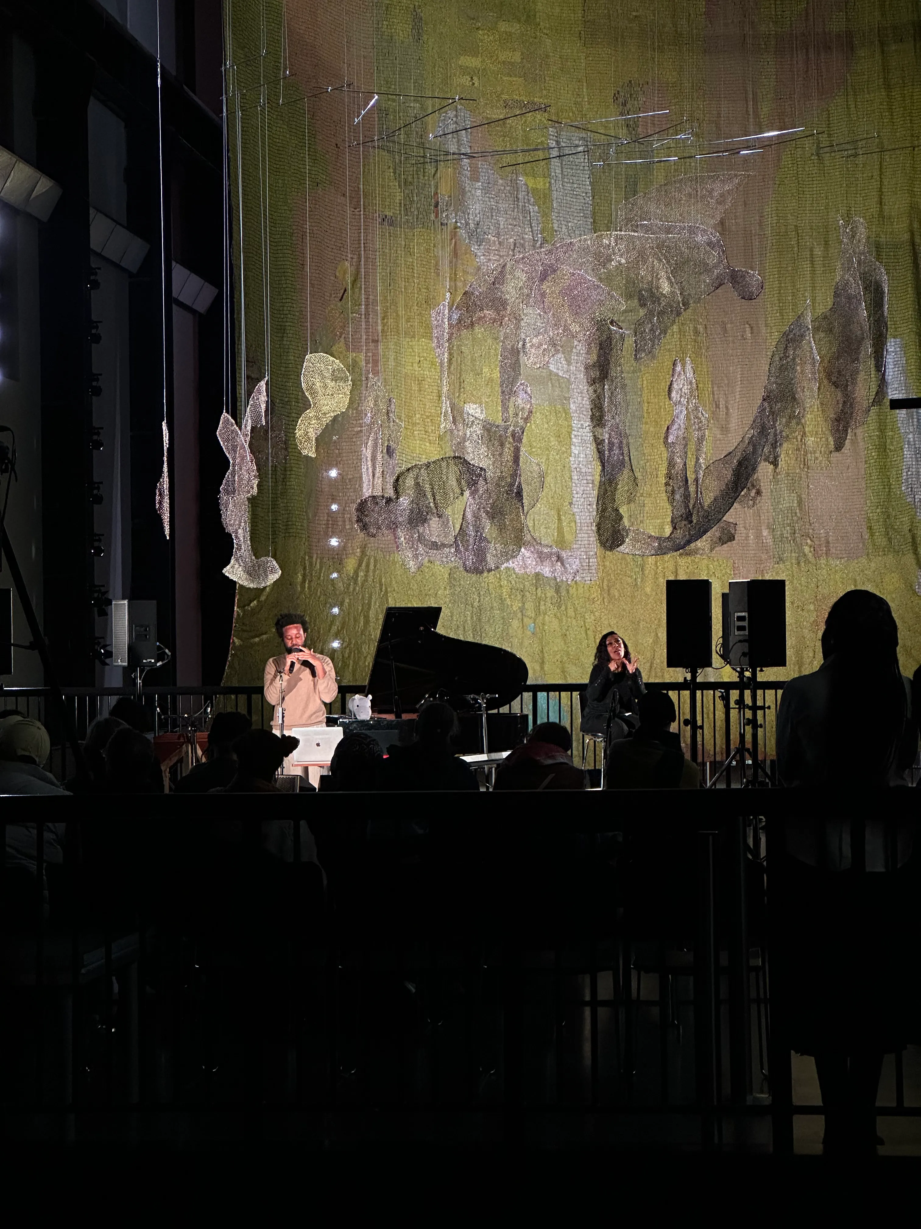 A photograph of JJJJJerome Ellis on stage performing, taken from someone in the crowd. He is singing into a microphone with a grand piano and various other instruments around him. In the backrgound is a massive gold-yellow sculpture constructed from glittering materials.