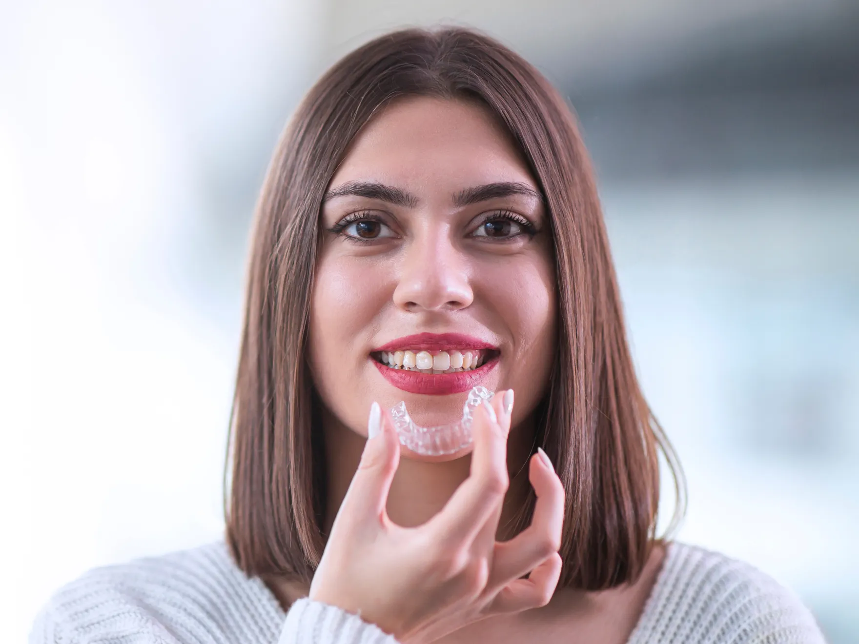 Smiling woman with brown hair holding a clear dental aligner near her mouth.