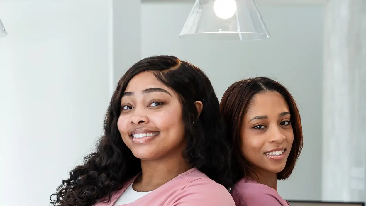 Two smiling women in pink uniforms standing back to back in a bright, modern indoor space.