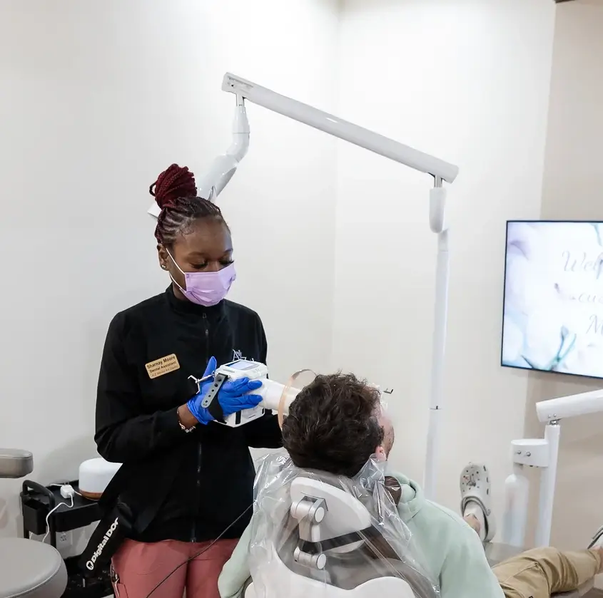 Dental assistant wearing mask and gloves takes X-ray of patient seated in dental chair.