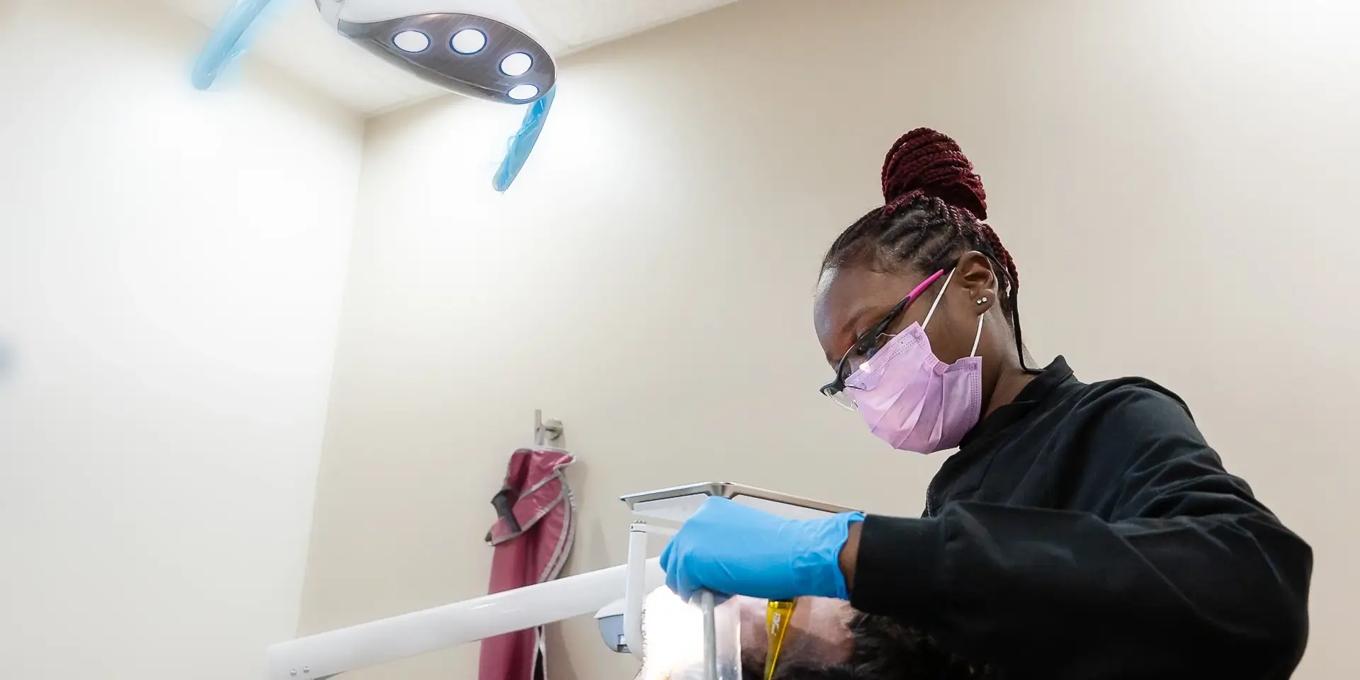 Dental professional wearing gloves and mask working on a patient's teeth under a bright lamp.