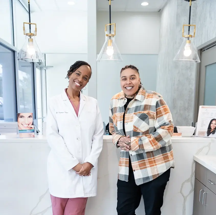 Smiling doctor in white coat and patient in plaid jacket stand together in a modern dental office.
