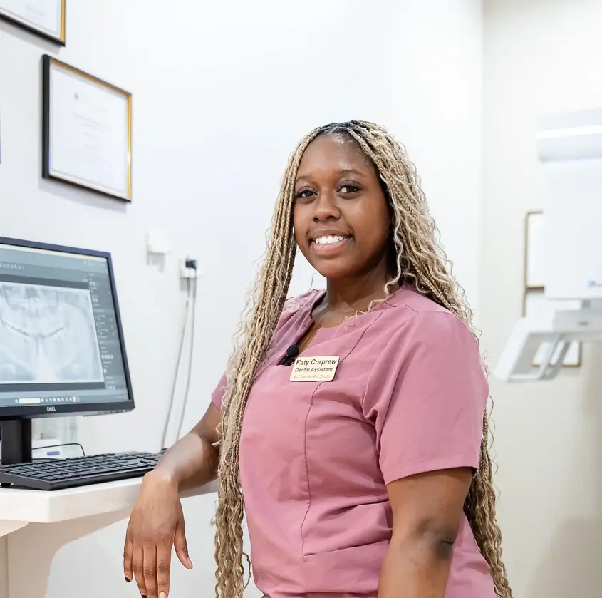 Smiling dental assistant in pink scrubs stands by a computer showing a dental X-ray image.