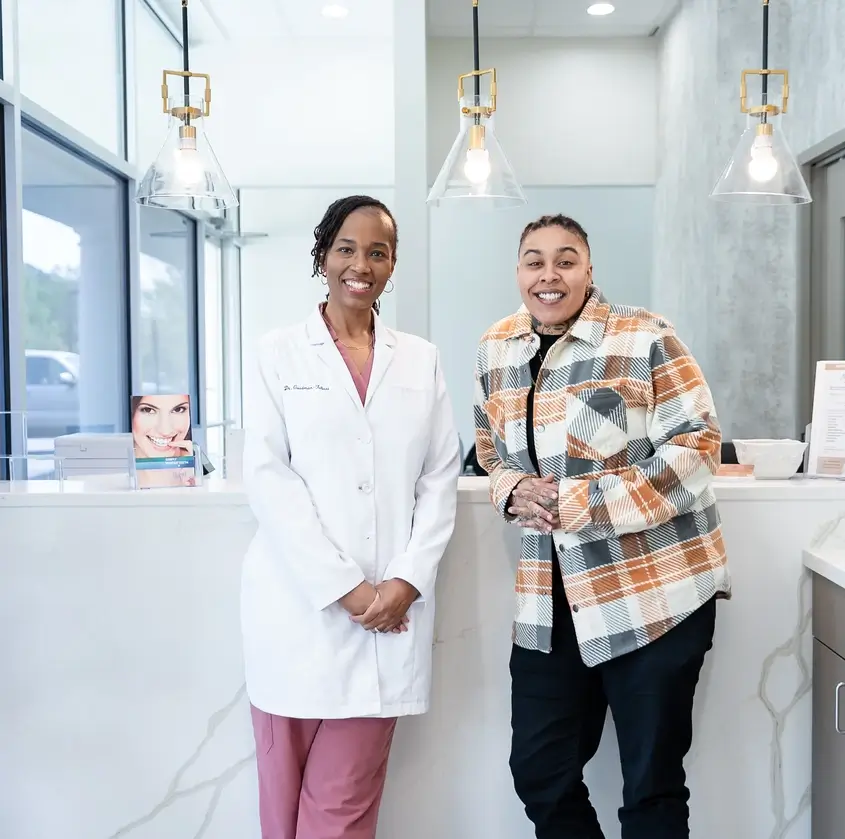 Smiling female doctor in white coat stands next to a casually dressed smiling person at reception desk.