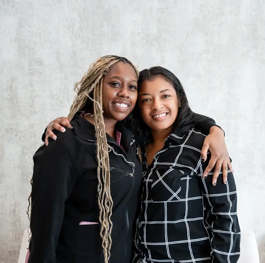 Two women smiling and hugging, one with long braided hair, the other wearing a black and white checkered shirt.
