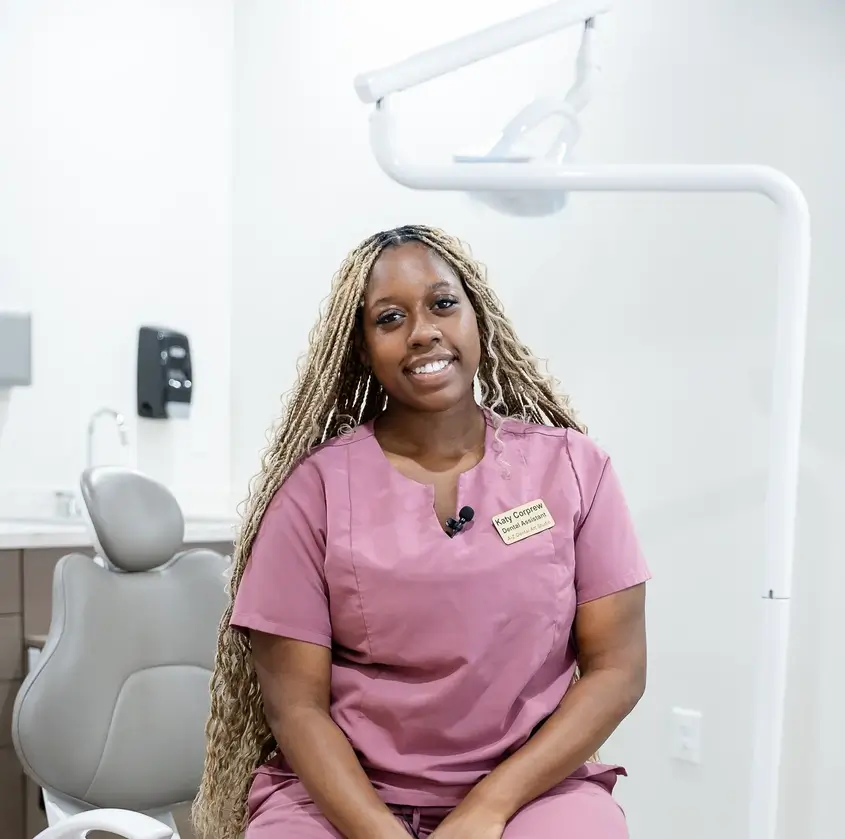 Woman in pink scrubs sitting in a dental office with a dental chair and equipment behind her.