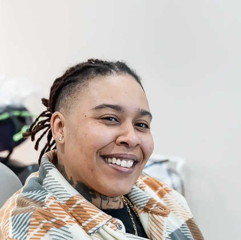 Person with short dreadlocks wearing a plaid jacket and a gold chain smiling indoors.