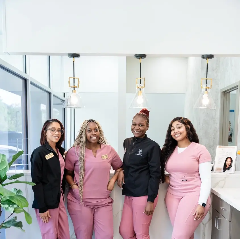 Four women in pink medical scrubs and black jackets smiling inside a bright clinic reception area.