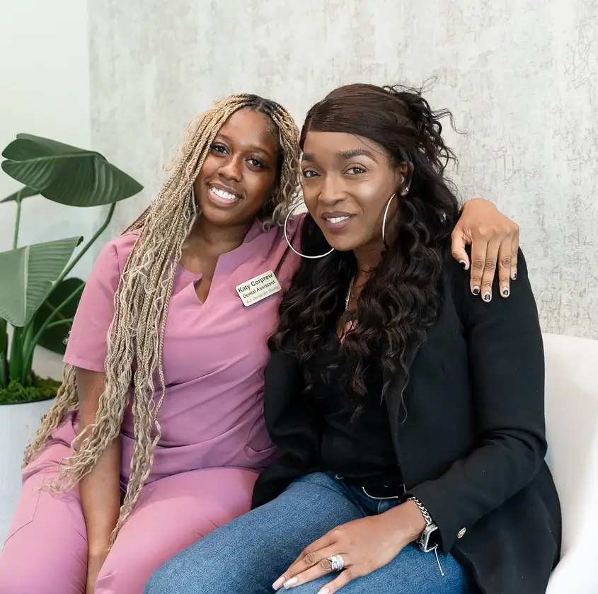 Two women sitting close together smiling; one in pink scrubs with a name tag, the other in black jacket and jeans.