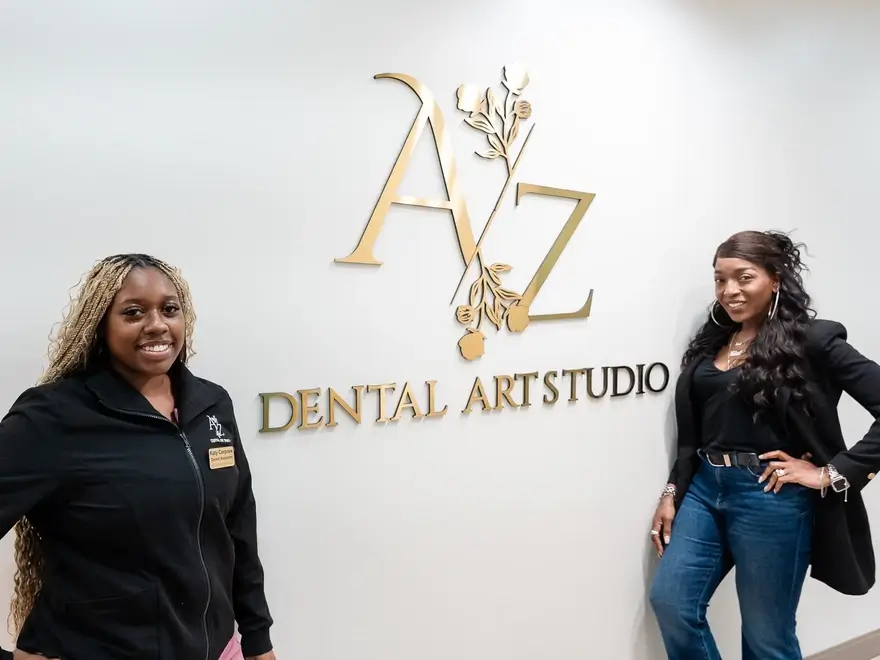 Two women standing and smiling in front of a wall with a gold sign reading AZ Dental Art Studio.