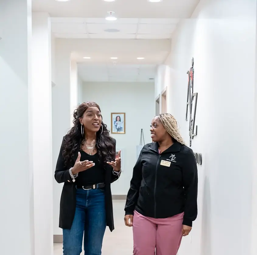 Two women walking and talking in a bright hallway with white walls and ceiling lights.