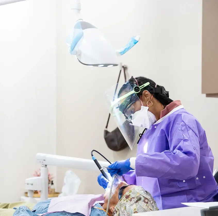 Dentist in purple gown and face shield treating patient lying in dental chair under overhead light.