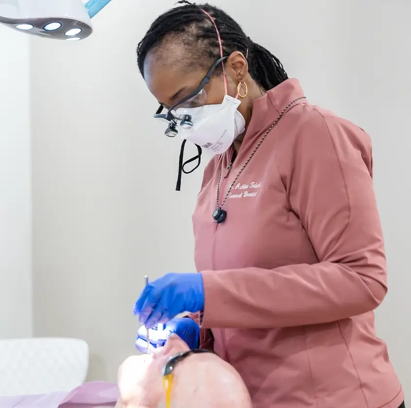 Dentist wearing mask and gloves examining a patient's mouth under dental light.