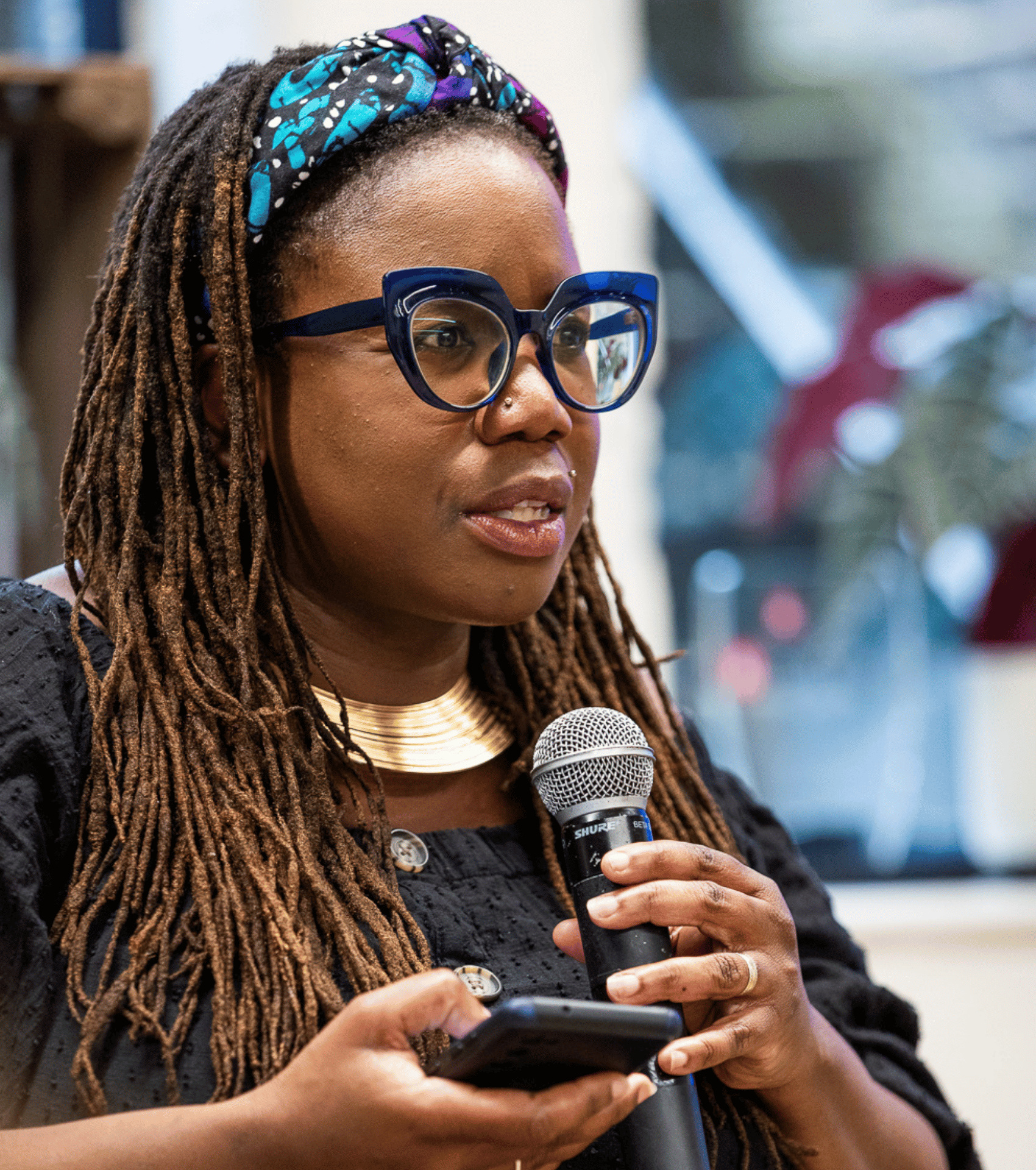 Collette speaking into a handheld microphone while holding a phone, wearing blue glasses, a patterned headband, and a gold necklace, addressing an audience at an indoor event.