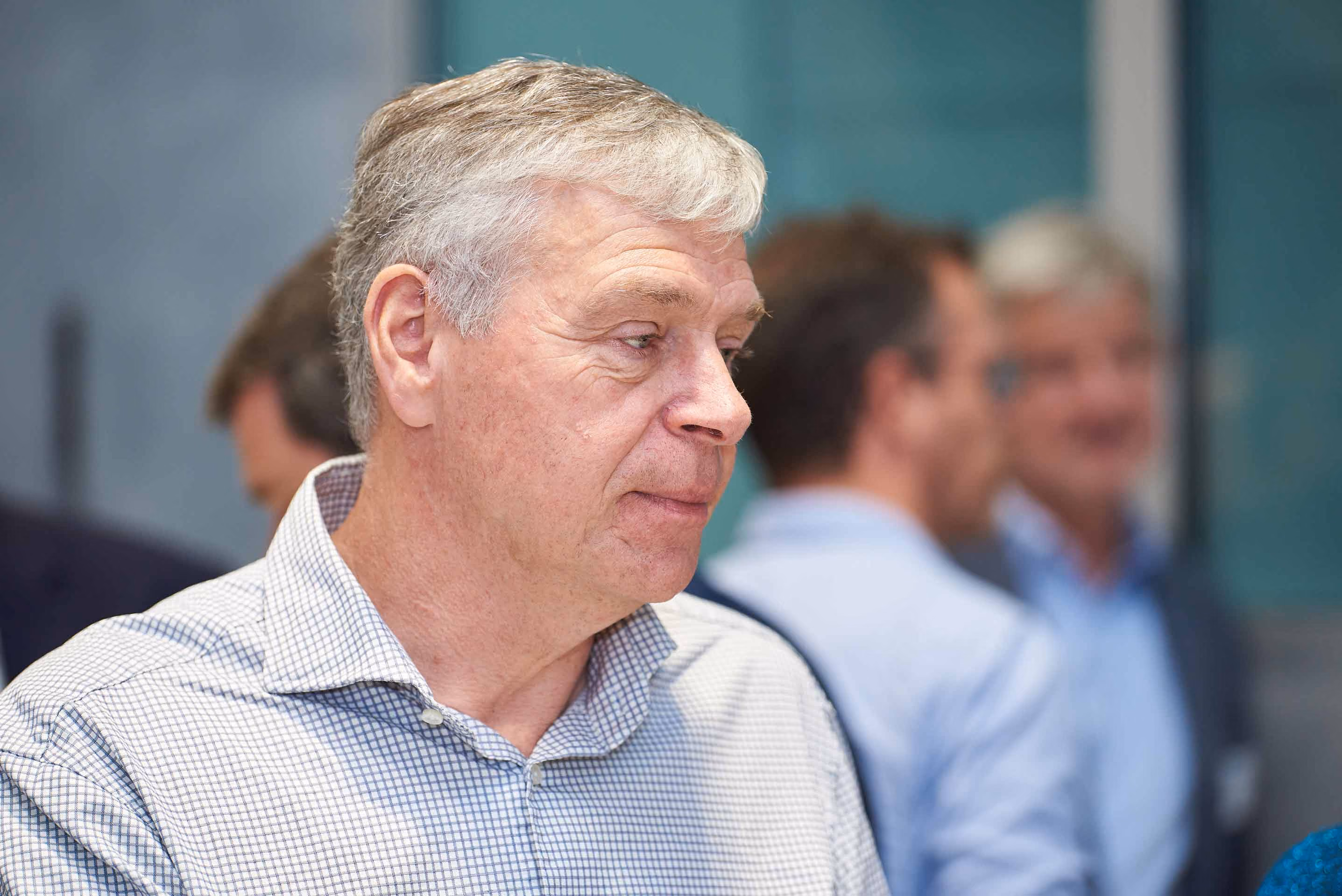 Close-up of an older man with gray hair wearing a checkered shirt, with two blurred men in the background engaged in conversation.