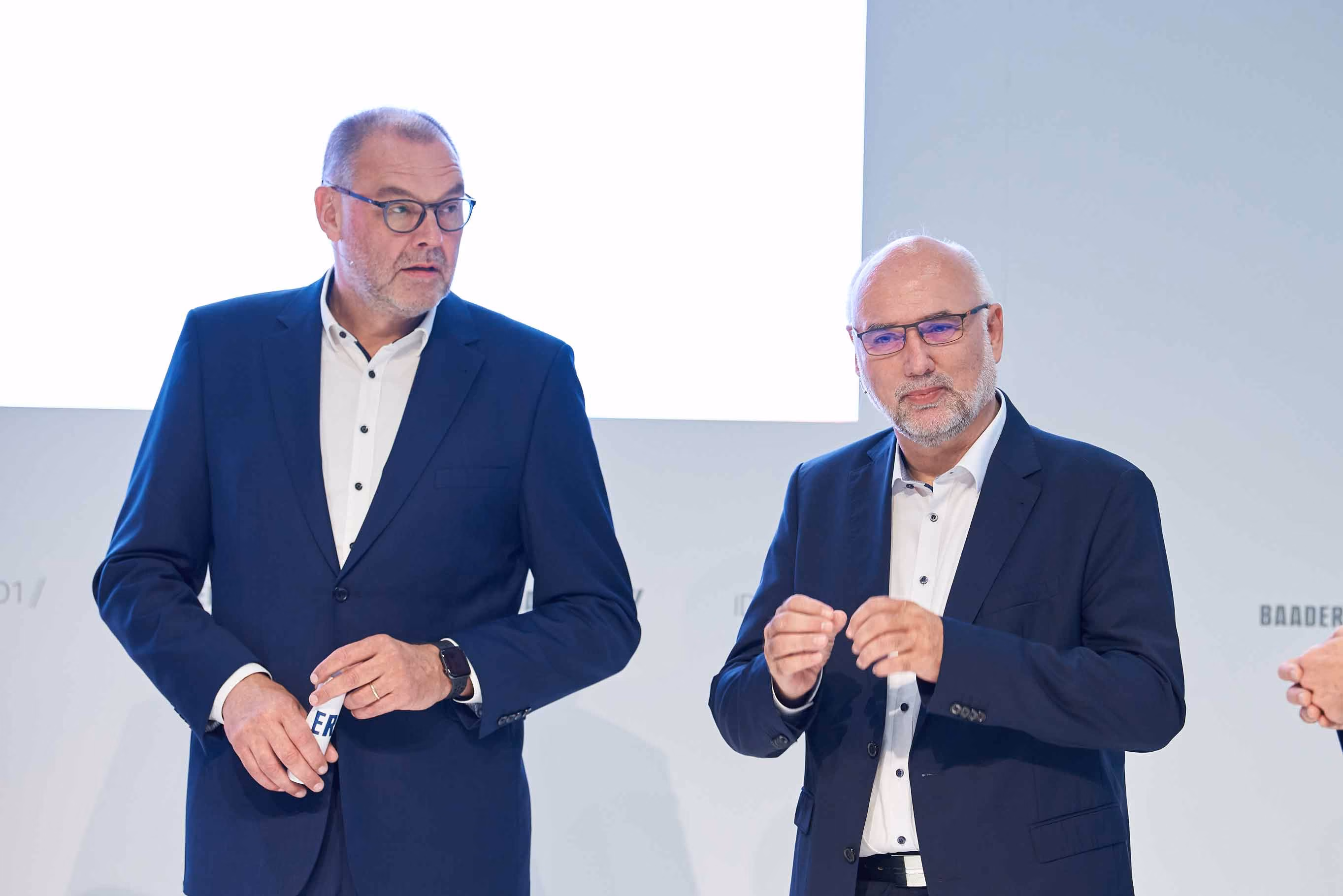 Two middle-aged men in dark blue suits and glasses speaking or presenting on a stage with a white background.