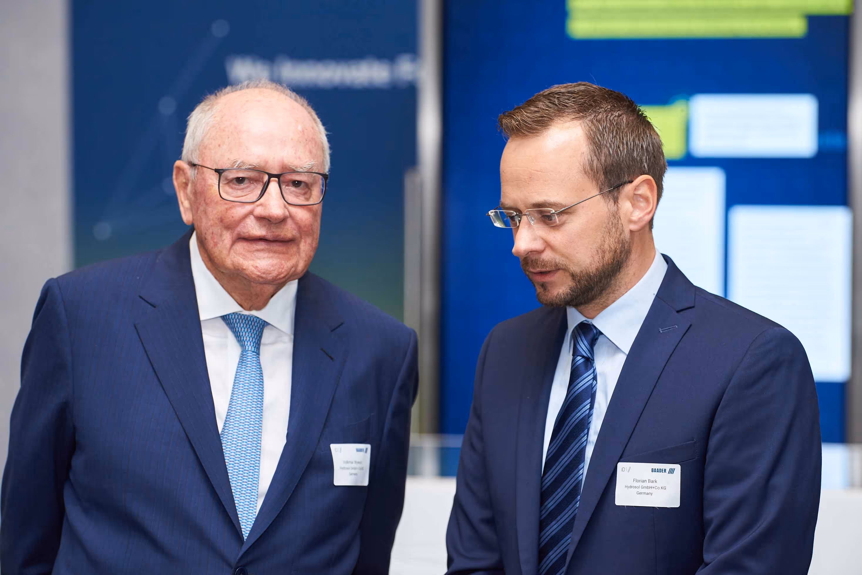 Two men in business suits with name tags standing and engaging in conversation at a formal event.