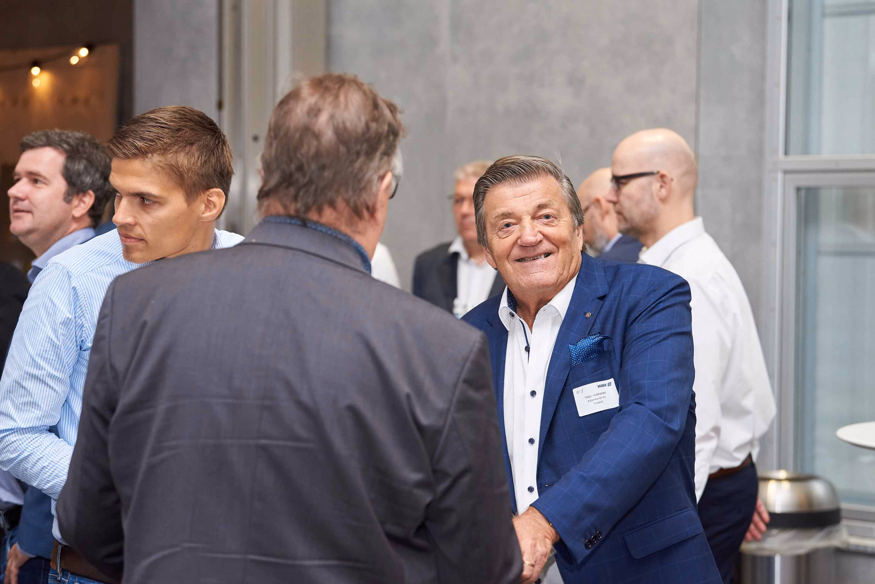 Older man in a blue suit smiling while shaking hands with another man at a professional networking event.