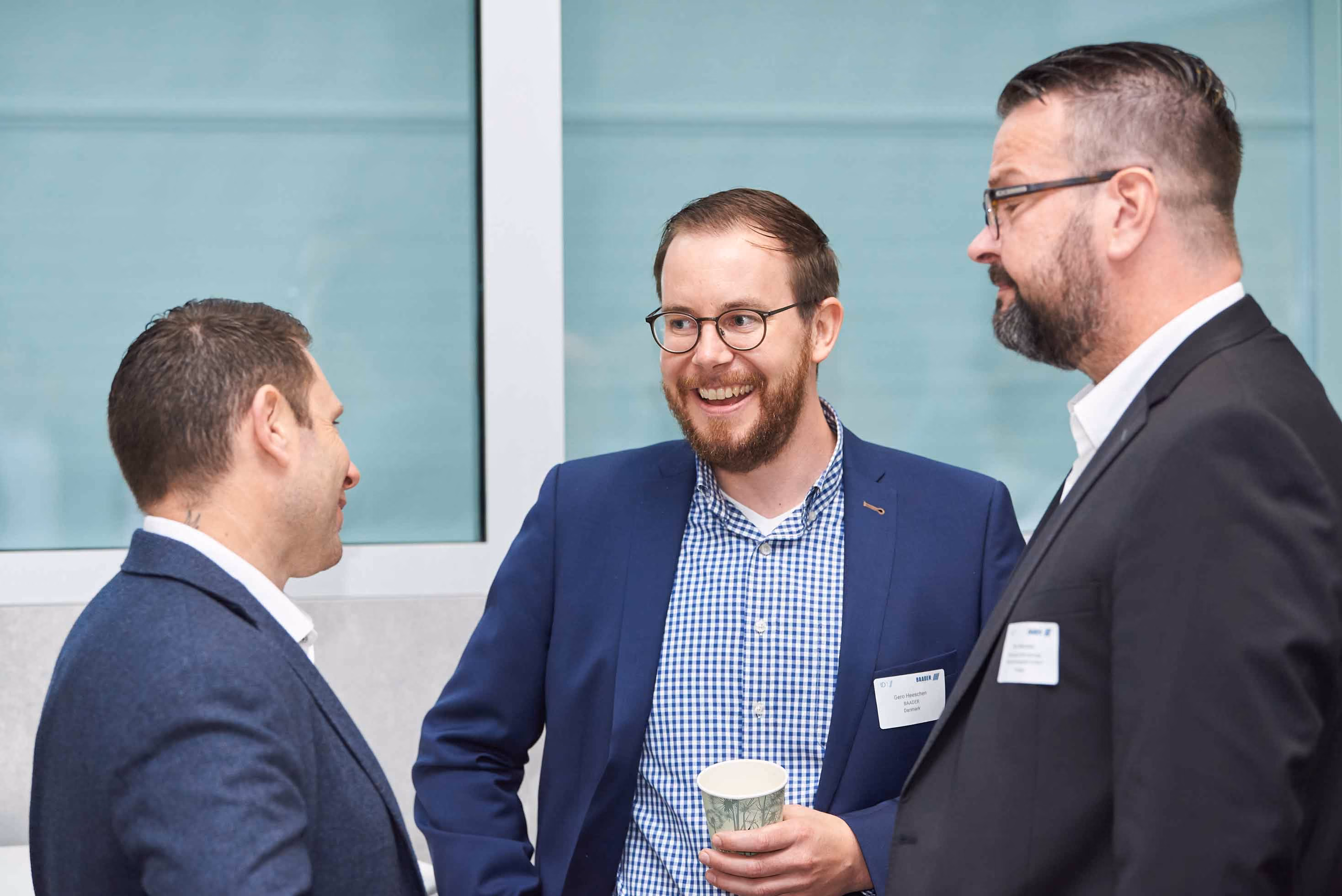 Three men wearing business attire engaged in conversation, with one holding a paper cup and smiling.