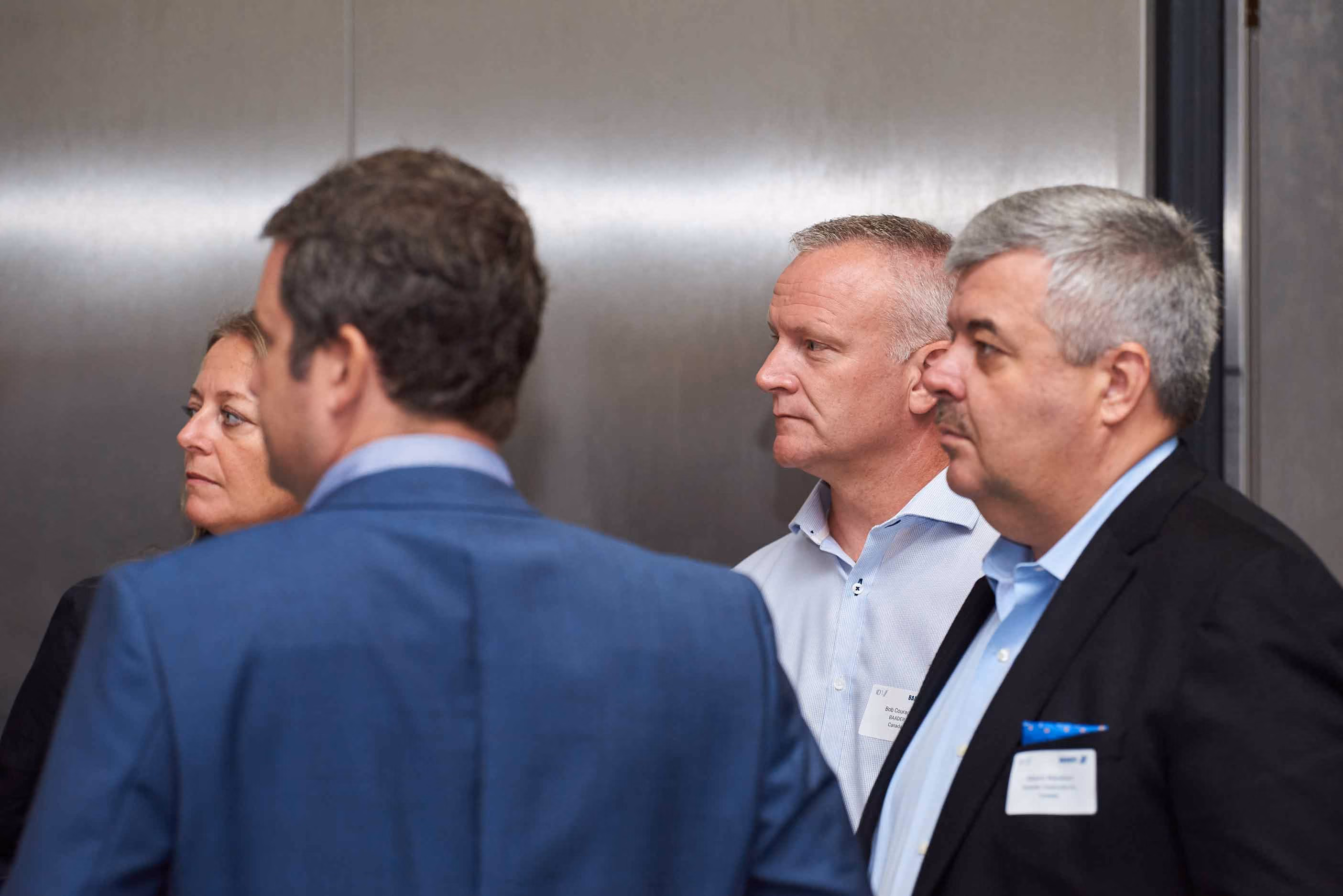 Four business professionals attentively listening, two men in front wearing suits and two individuals in the background with name tags.