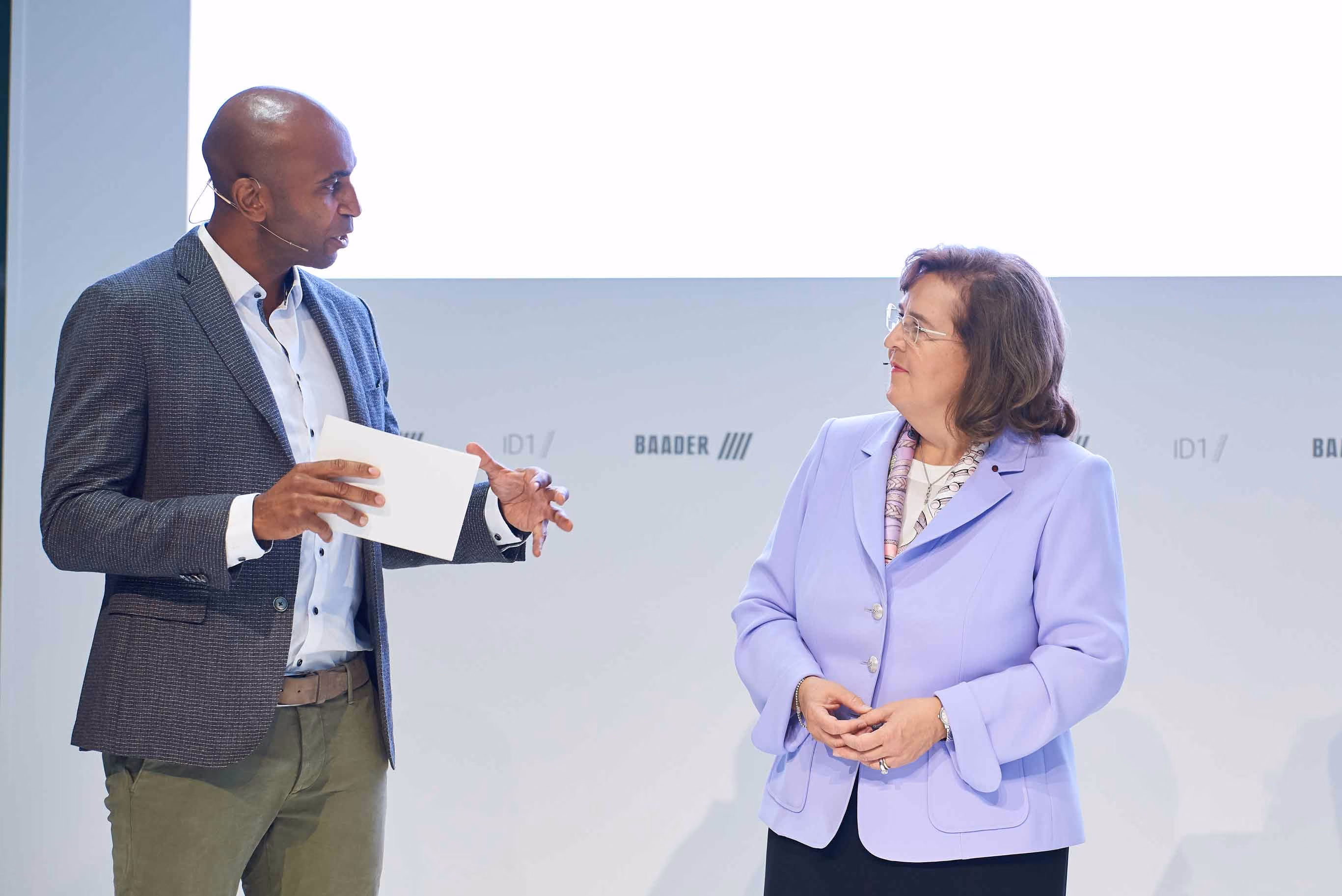 Two persons having a discussion on stage during an event, one holding a piece of paper.